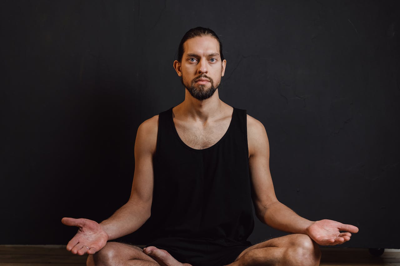 Bearded man meditating in a black tank top against a dark background, promoting peace and wellness.